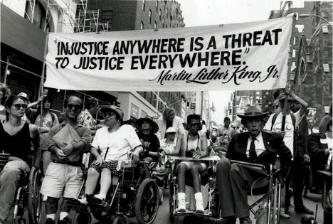 a black and white photograph of activists marching in New York City to demand passage of disability rights legislation. The diverse group includes several people with visible disabilities, and two activists hold a large banner that says “injustice everywhere is a threat to justice everywhere” and attributes the quote to Martin Luther King Jr.